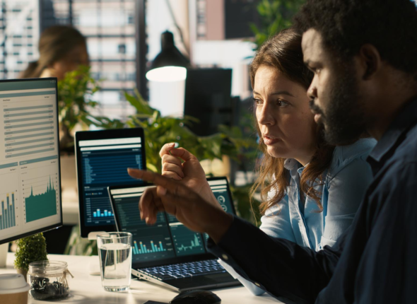 Two business professionals, a Black man and a White woman, review financial data on computer screens displaying charts and graphs, symbolizing data intelligence in sustainable finance.