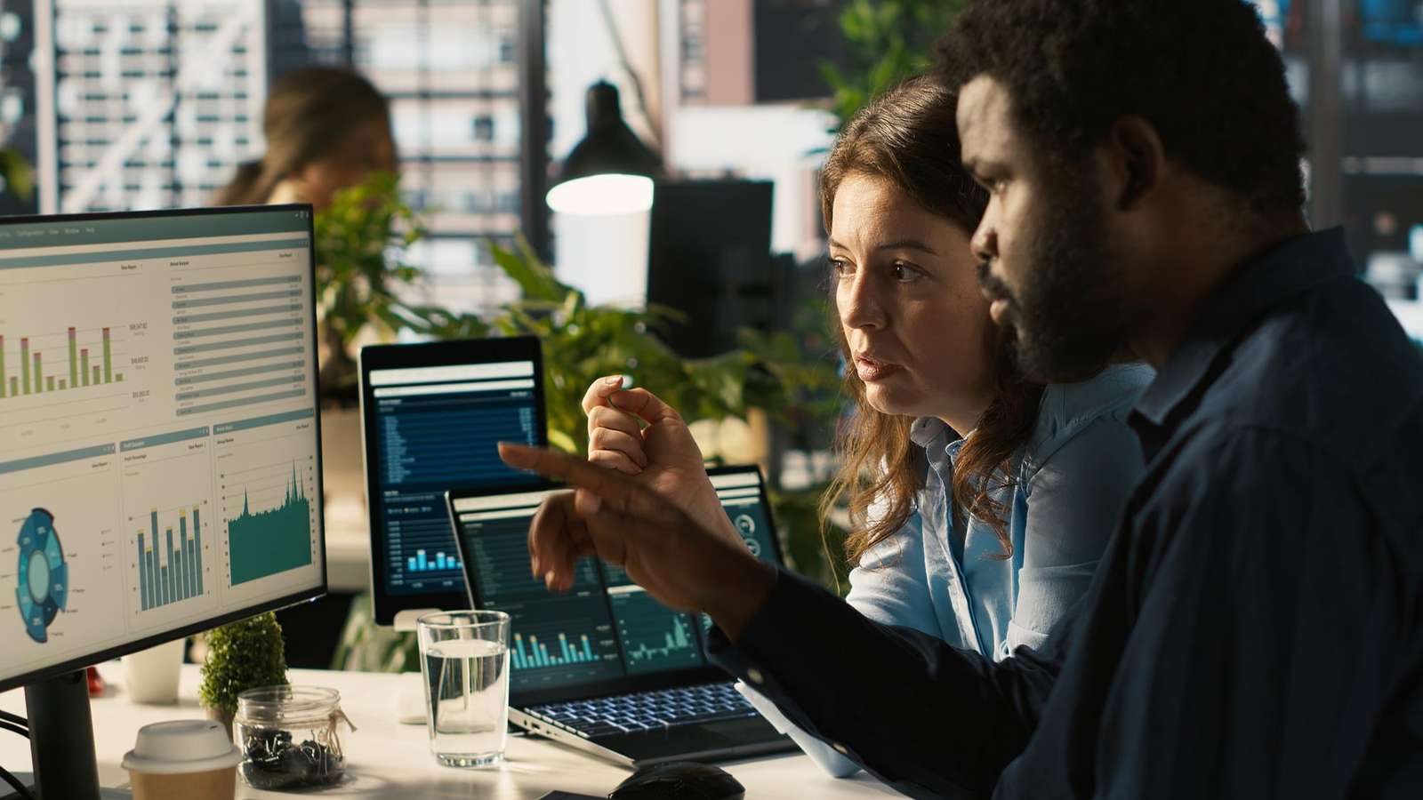 Two business professionals, a Black man and a White woman, review financial data on computer screens displaying charts and graphs, symbolizing data intelligence in sustainable finance.