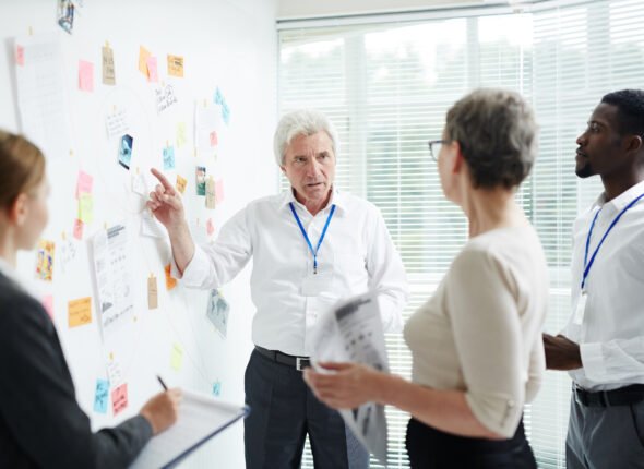 A diverse team of business professionals, including a senior white man, a white woman, and a black man, are gathered around a whiteboard covered in sticky notes, documents, and red string connections. The senior man is pointing at the whiteboard, appearing to explain a complex strategy.