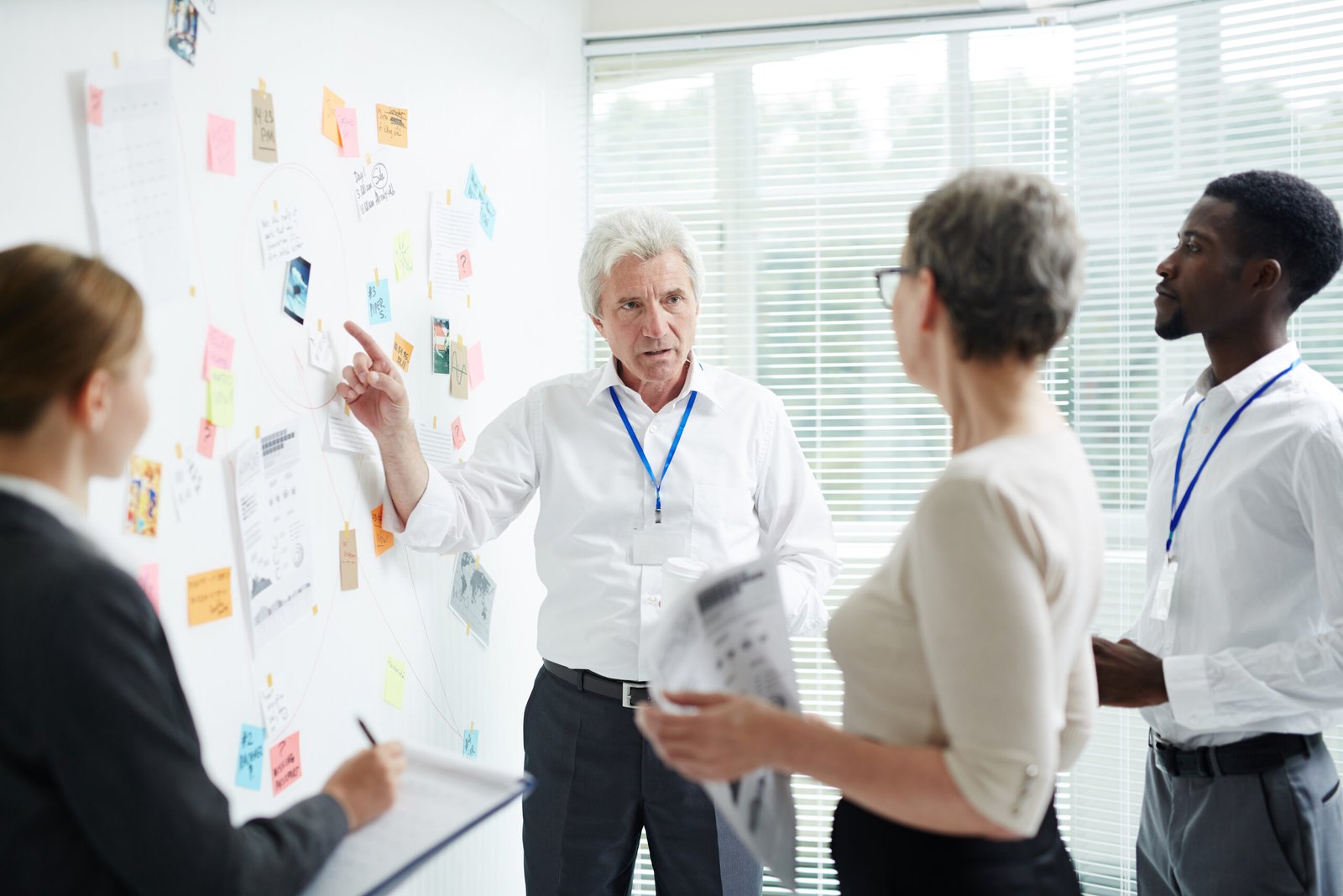 A diverse team of business professionals, including a senior white man, a white woman, and a black man, are gathered around a whiteboard covered in sticky notes, documents, and red string connections. The senior man is pointing at the whiteboard, appearing to explain a complex strategy.